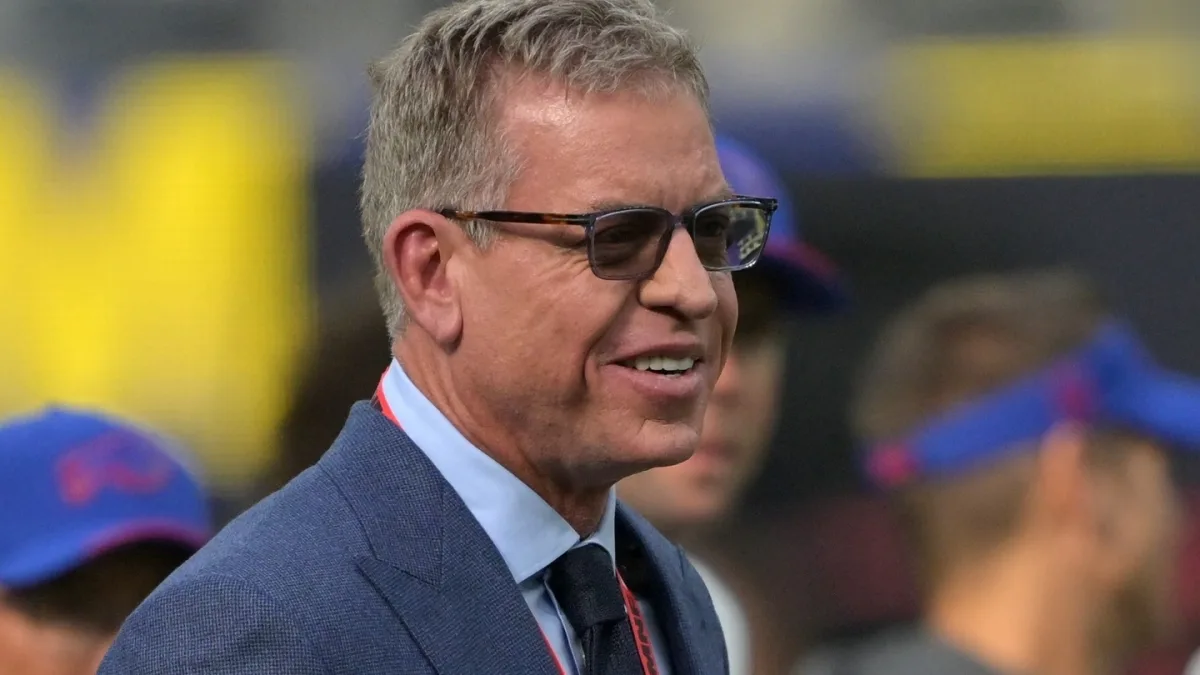 Troy Aikman in a suit and tie, smiling and holding a microphone